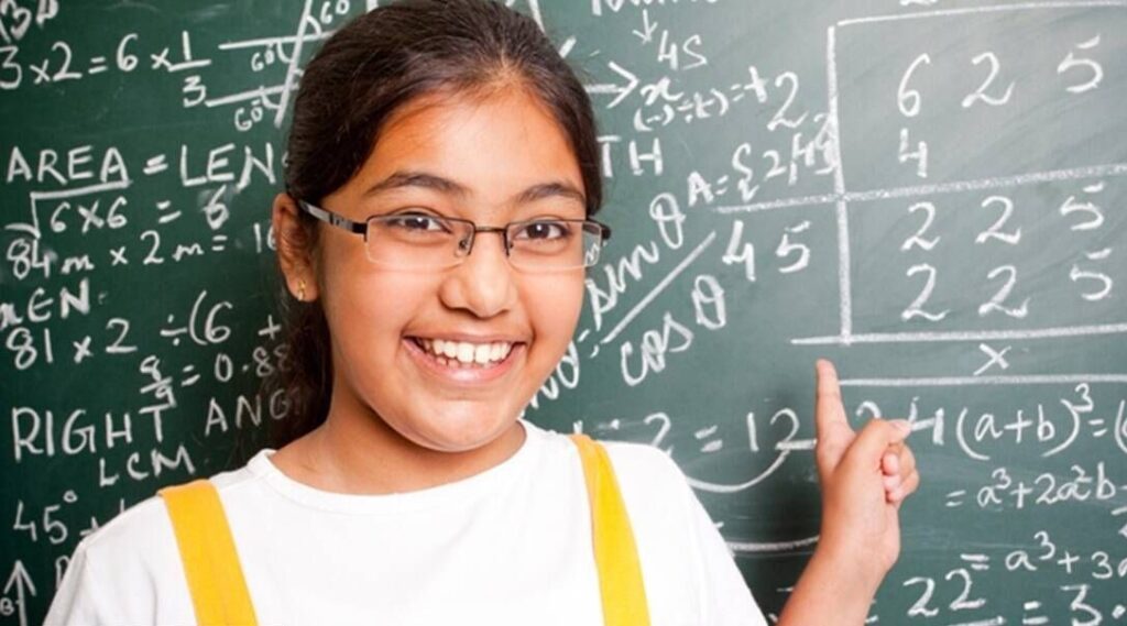 Young girl smiling and pointing at a chalkboard filled with mathematical equations.